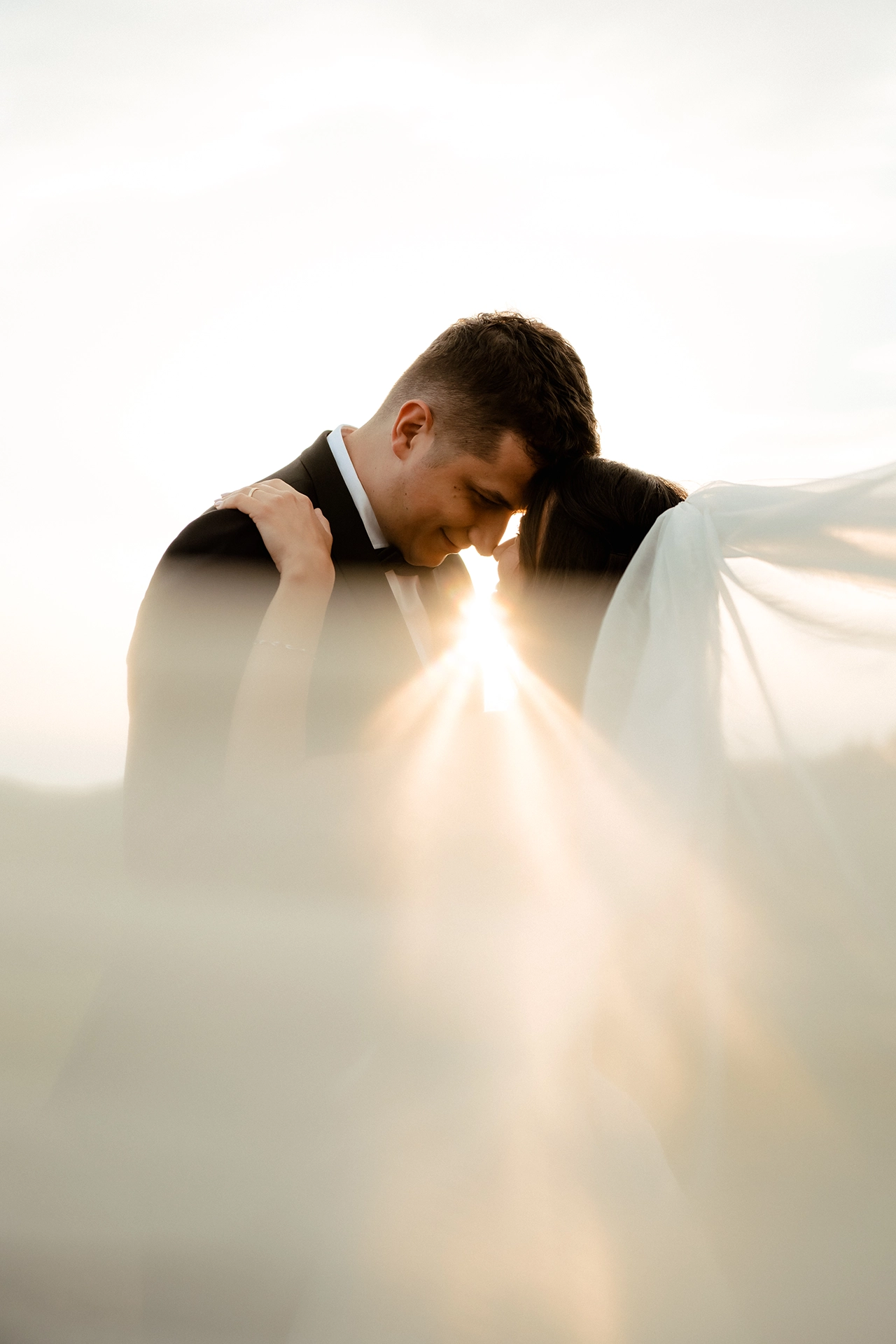 Bride and groom touching foreheads at sunset with sunlight between them and her veil blowing in the wind