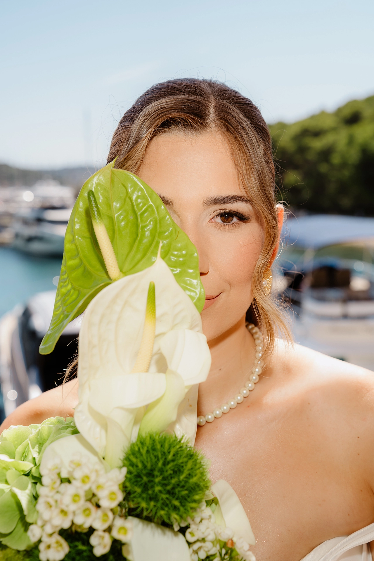 Editorial portrait of a bride in a pearl necklace hiding half her face behind a green and white anthurium bouquet at Marina Frapa in Rogoznica, Croatia
