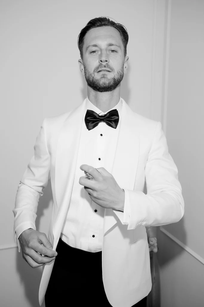 Black and white flash portrait of a groom in a white tuxedo jacket and black bow tie spraying cologne while getting ready in Opatija, Croatia