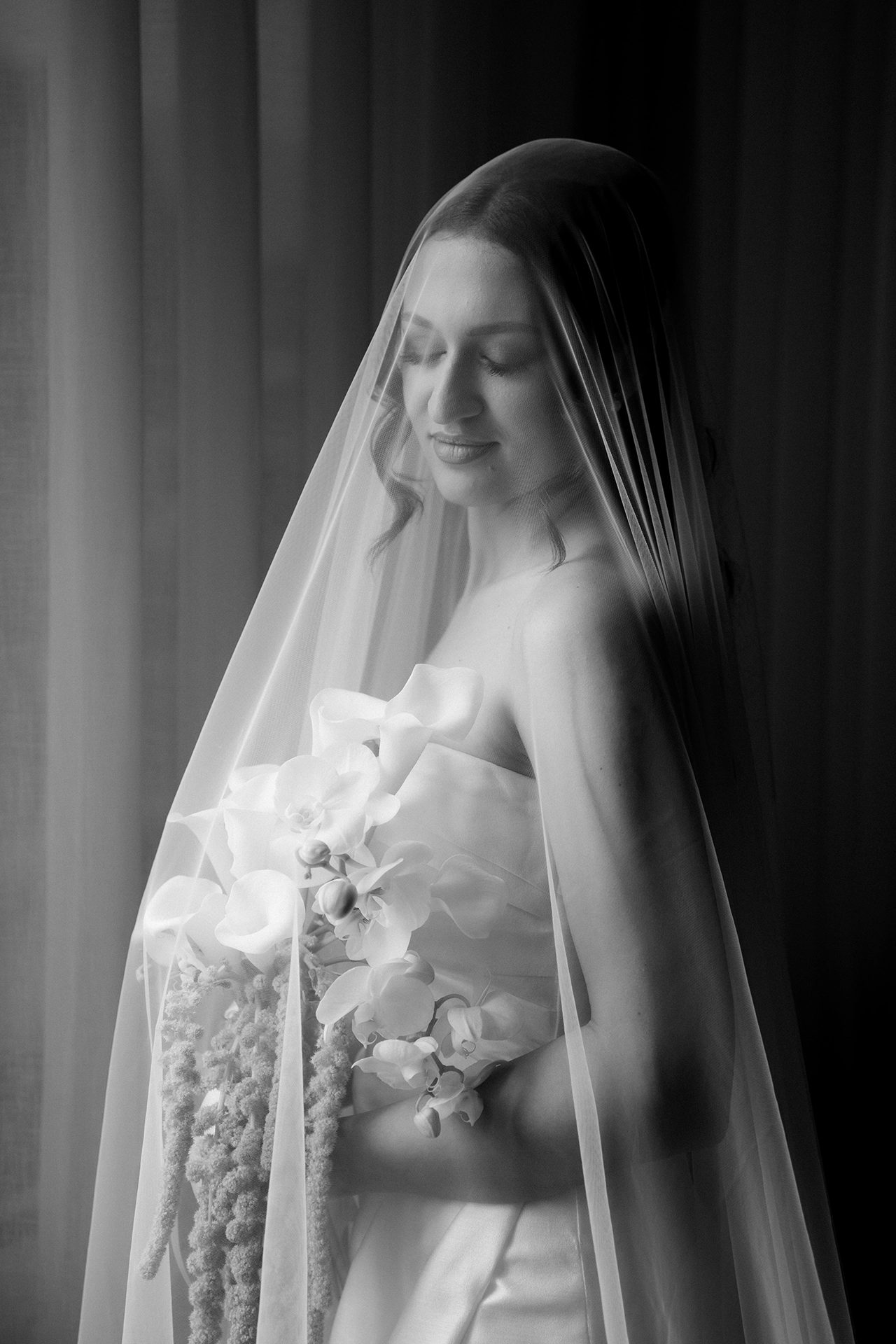 Black and white portrait of a smiling bride under a long sheer veil holding a cascading bouquet of calla lilies, orchids and amaranthus by the window