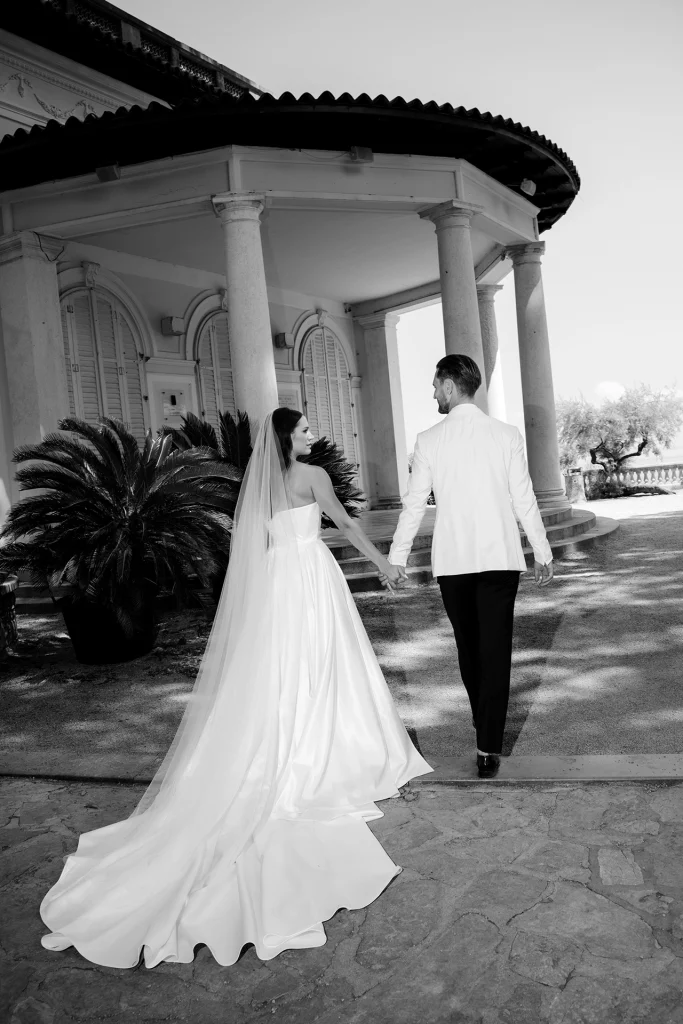 Black and white photo of a bride and groom walking hand in hand toward a columned villa in Opatija, Croatia, her long veil and train trailing behind