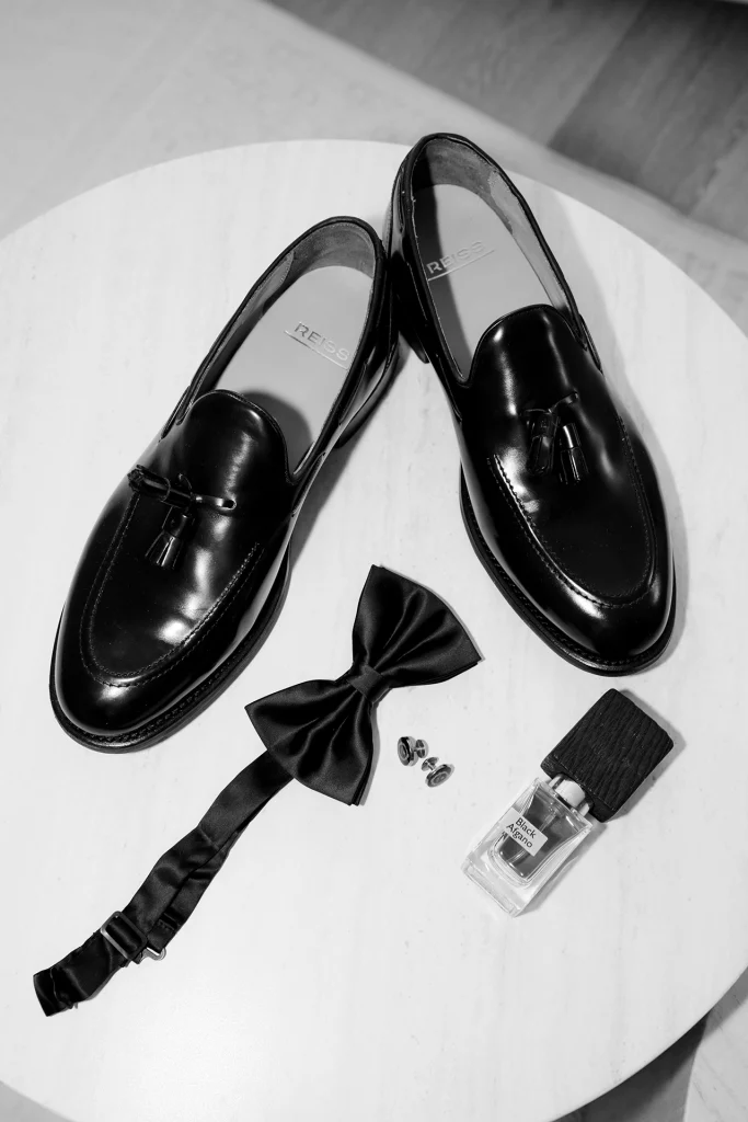 Black and white flat lay of groom’s details with black tassel loafers, a silk bow tie, cufflinks and cologne on a round white table