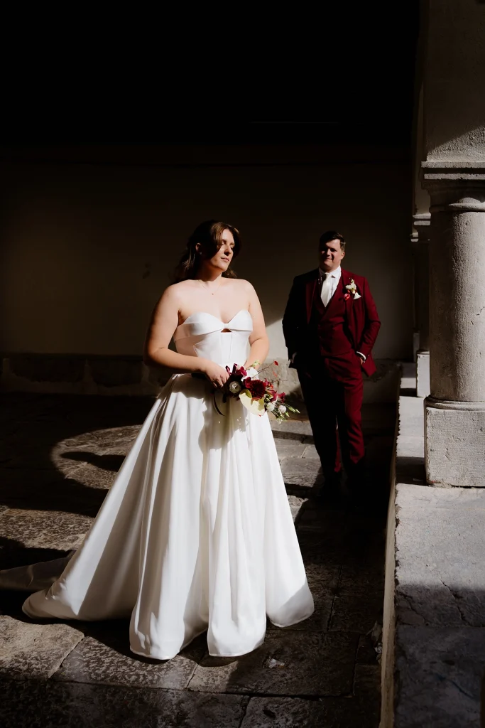 Bride in a strapless satin ball gown standing in dramatic sunlight in a stone cloister in Rijeka, Croatia, with the groom in a burgundy suit behind her
