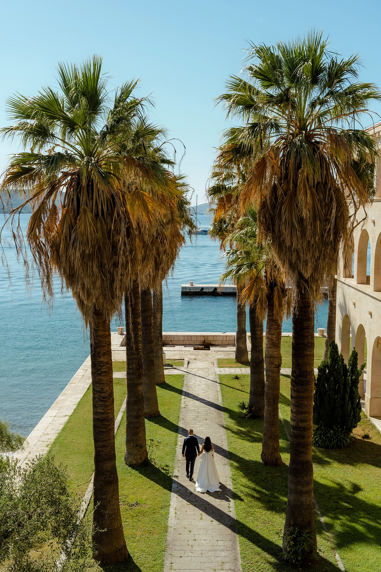 Bride and groom walking hand in hand down a palm-lined path toward the sea beside an arcaded stone building in Split, Croatia