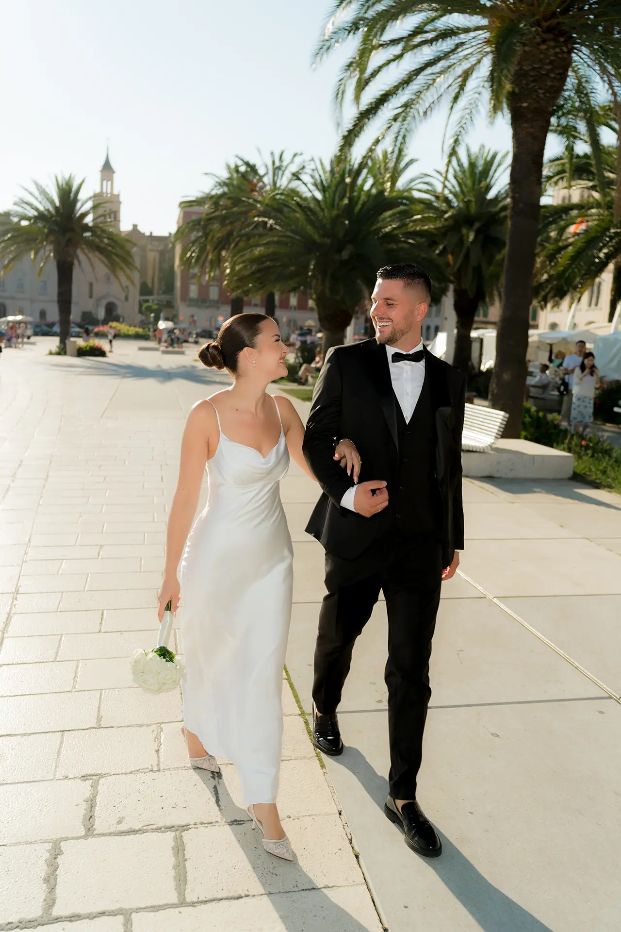 Bride in a satin slip dress and groom in a black tuxedo walking arm in arm along the palm-lined Riva promenade in Split, Croatia