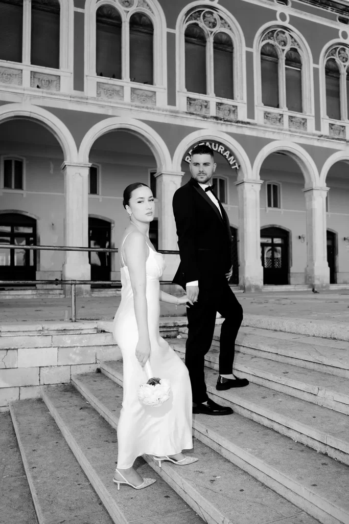 Black and white photo of a bride and groom walking up the steps in front of the Prokurative arcades on Republic Square in Split, Croatia