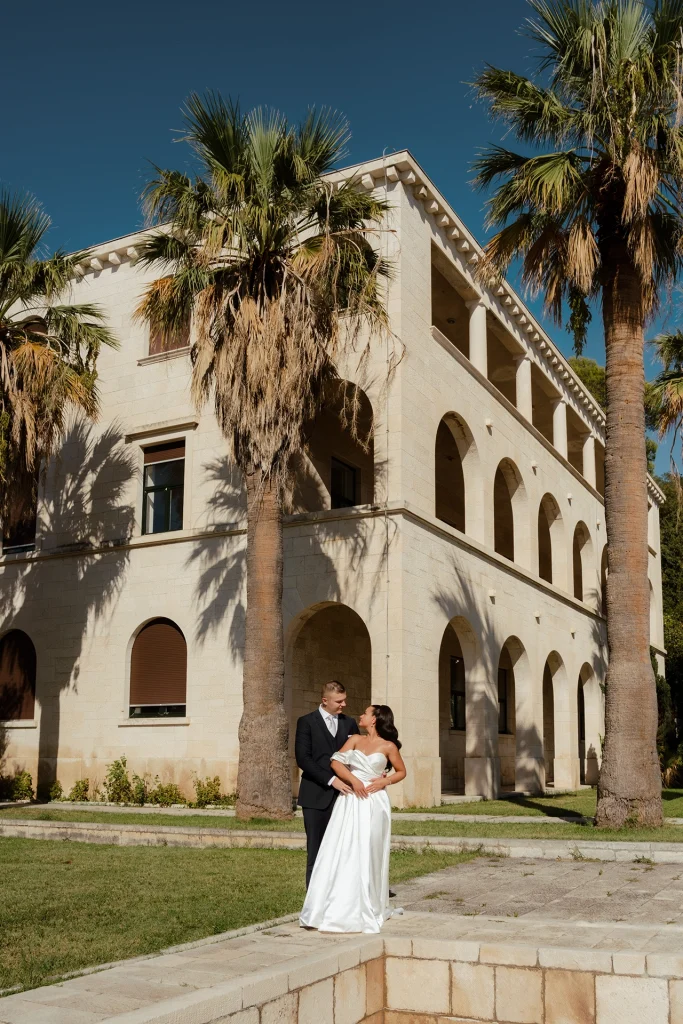 Bride and groom embracing in front of an arcaded stone building with tall palm trees in Split, Croatia