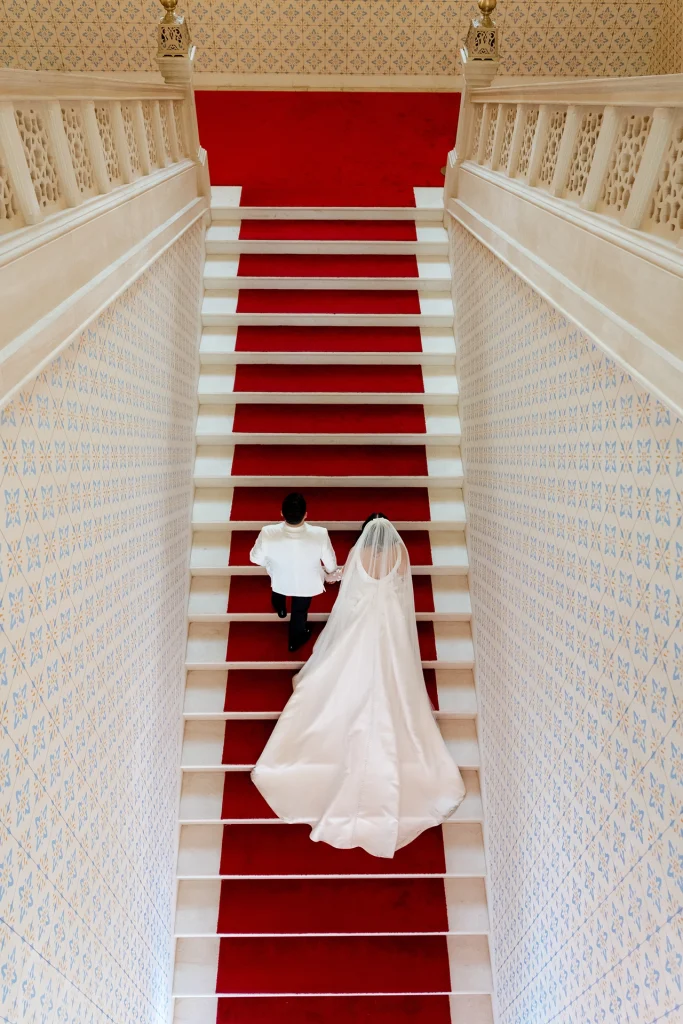 Overhead photo of a bride and groom walking up the red-carpeted staircase of Sarajevo City Hall (Vijećnica), her long veil and train trailing behind