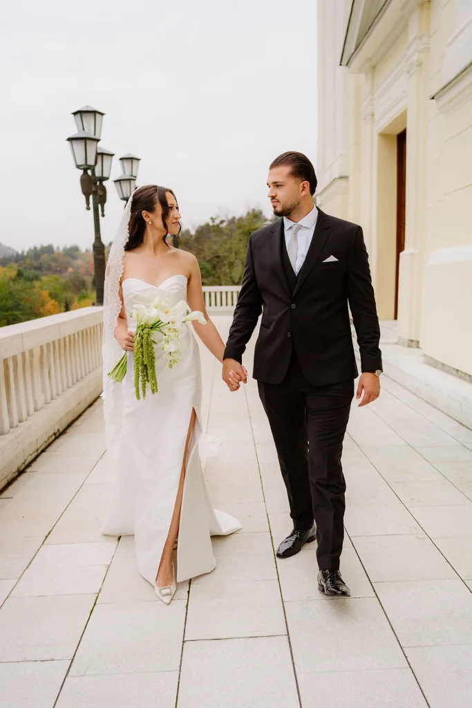 Bride and groom walking hand in hand on a white terrace with vintage lamp posts, the bride holding calla lilies and green amaranthus