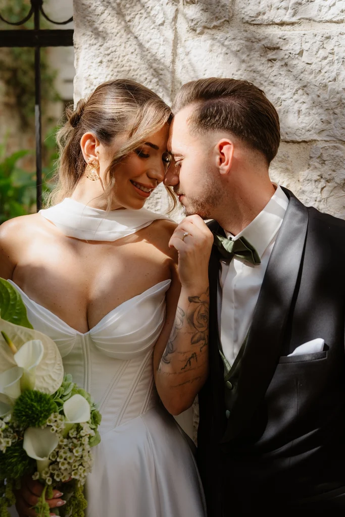 Bride and groom touching foreheads against a sunlit stone wall, the bride holding a white anthurium and calla lily bouquet