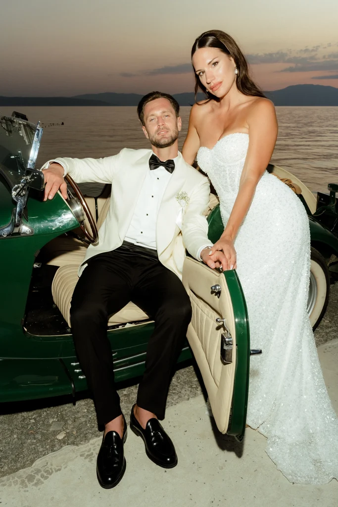 Groom in a white tuxedo jacket sitting in a vintage green convertible with the bride in a sequin gown leaning on the door by the sea at dusk in Opatija (Abbazia), Croatia